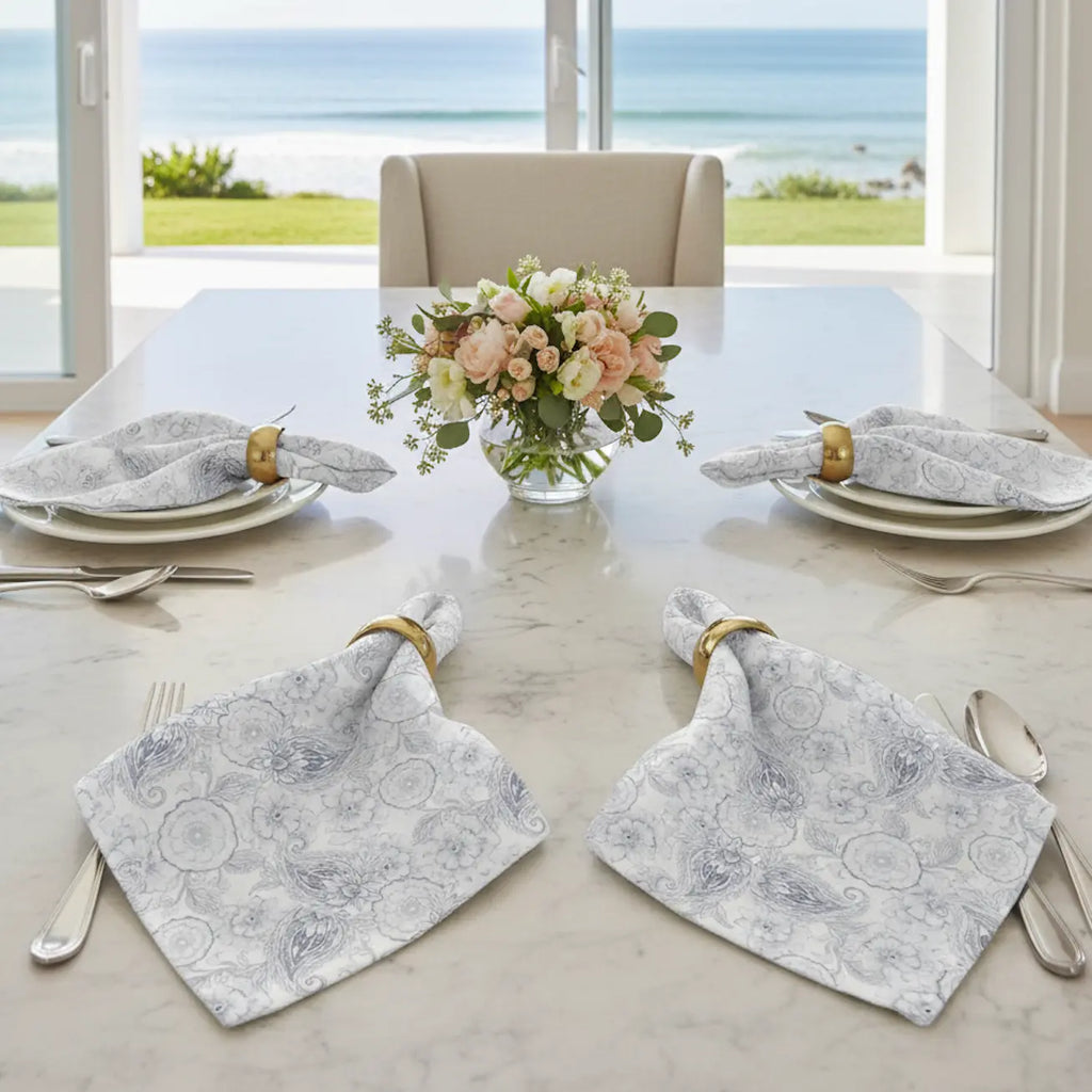 Blue paisley floral cloth napkins set of four with gold napkin rings on a marble table by seaside window.