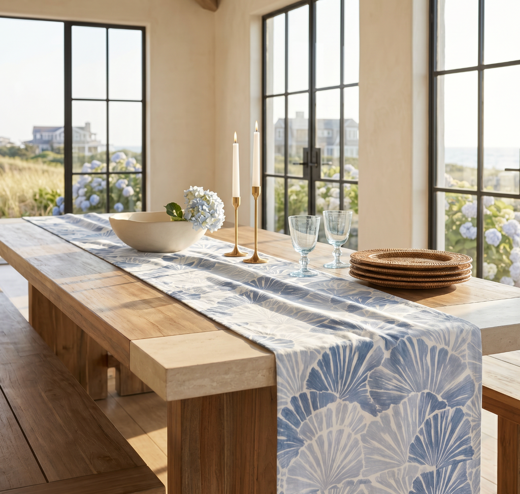 Blue and white coastal table runner with shell design on a long wood table set with woven chargers and linen napkins.