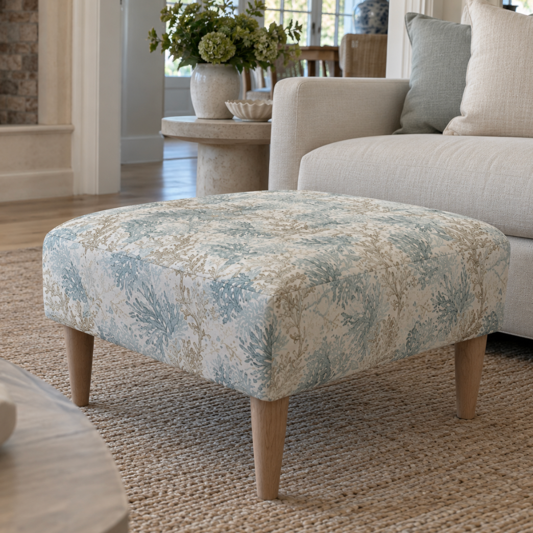 Square upholstered ottoman with blue and sage coral print fabric and tapered wood legs in a coastal-style living room