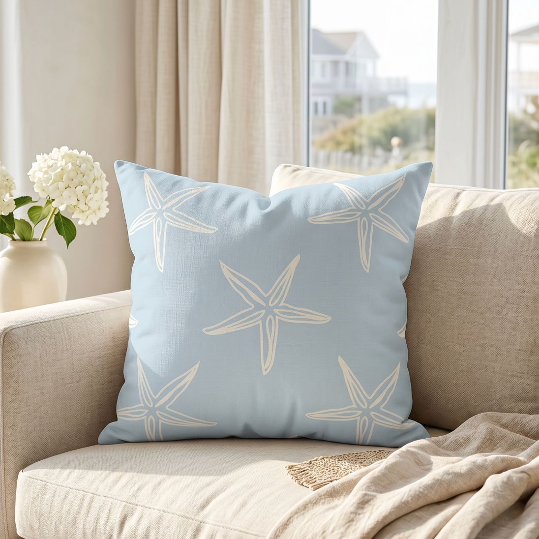 Light blue and white starfish print decorative pillow styled on a cream couch in a beach house living room.”