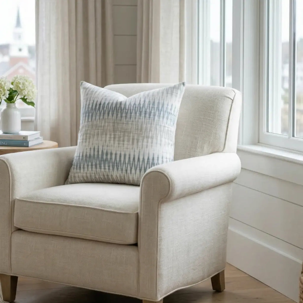 Stonewash Ikat Throw Pillow on a beige armchair by a sunlit window, blue and neutral ikat pattern.
