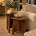 Organic modern wooden accent table with a sculptural silhouette styled beside an upholstered chair in a neutral living space.