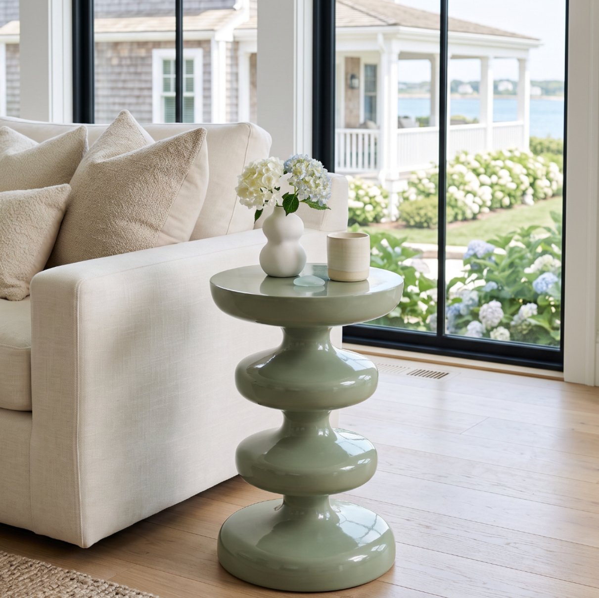 Glossy sage green metal side table with a round top and sculptural stacked pedestal base beside a white sofa.