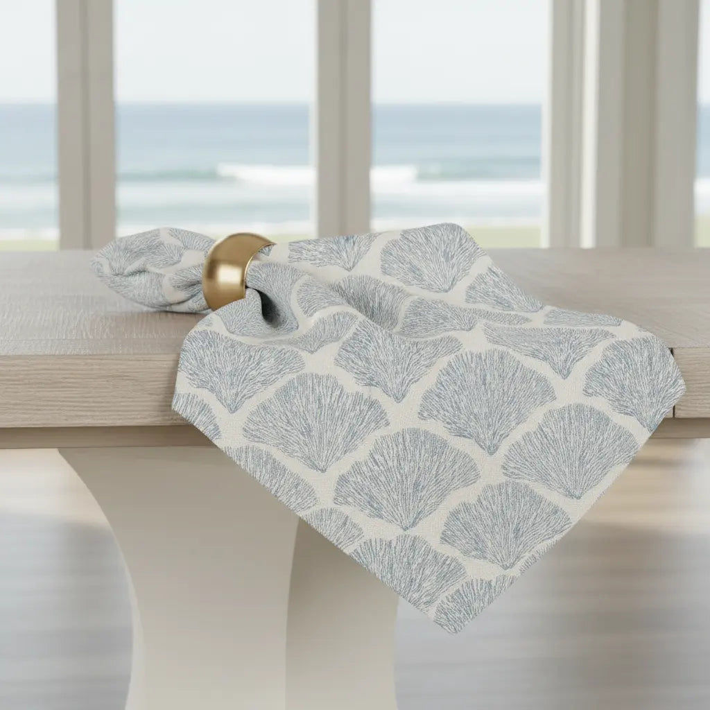 Bar Harbor Coral Linen Napkins, set of 4, blue seashell pattern draped over a light wood table with brass napkin ring.