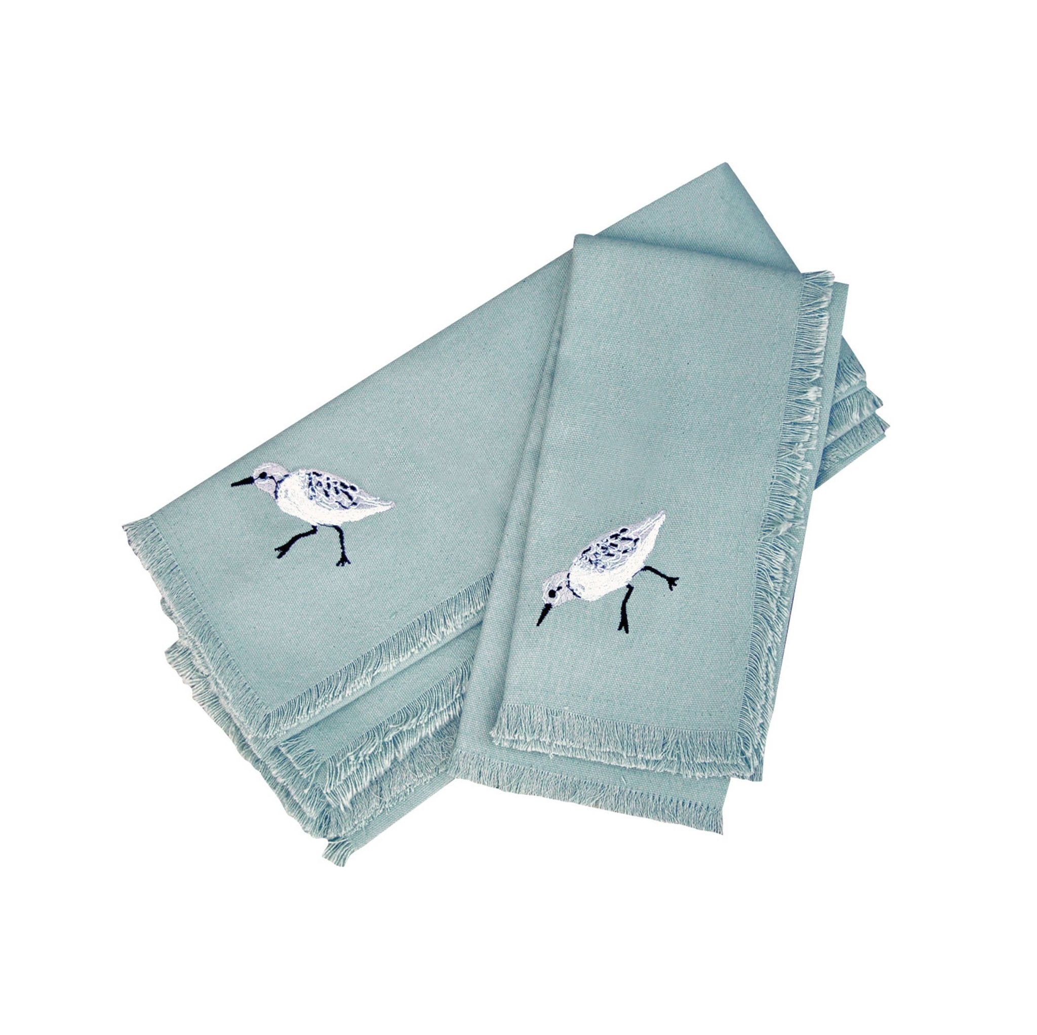Coastal embroidered bird napkins arranged with rattan rings, white plates, and ocean‑view table styling.