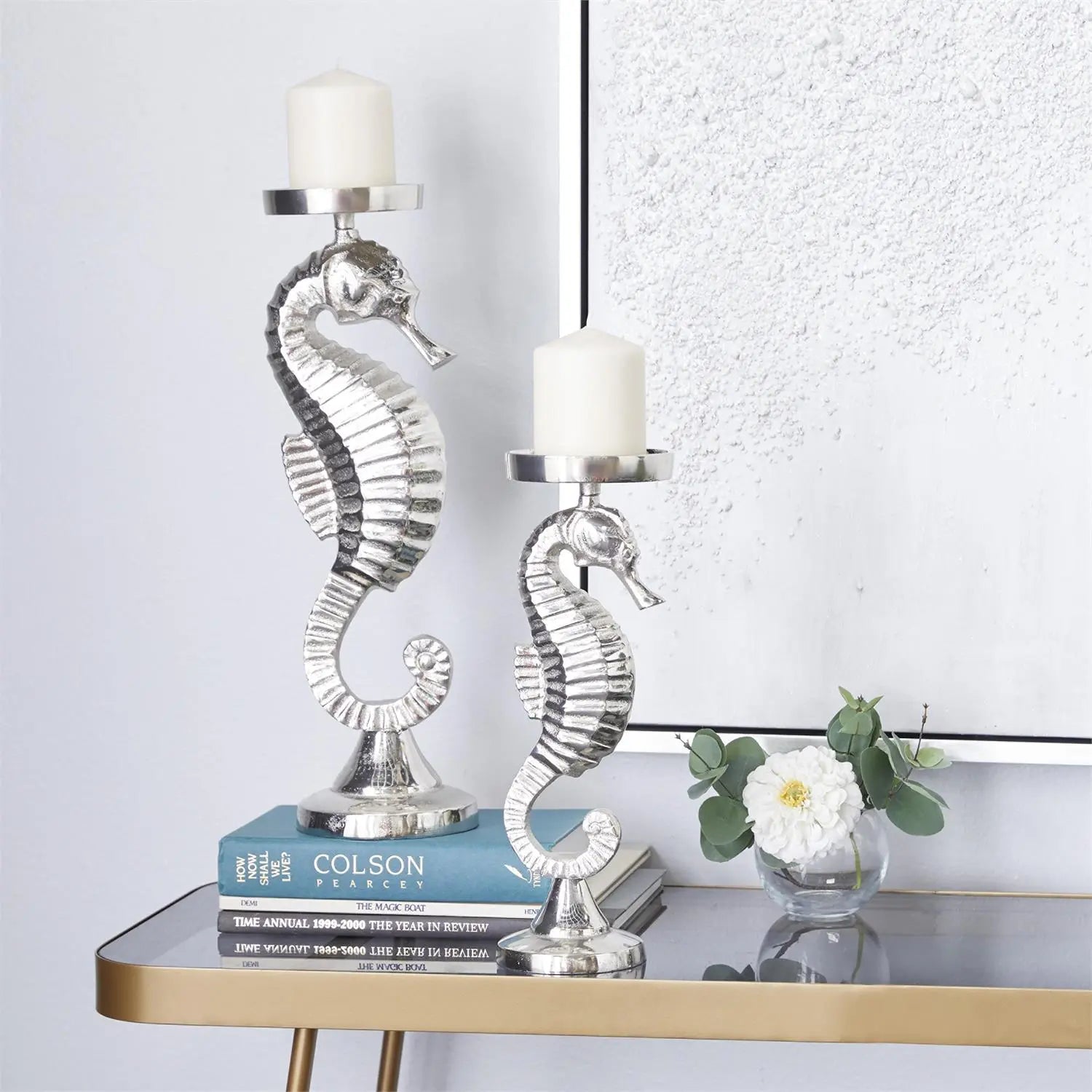 Polished silver seahorse candle holders with white candles on a glass-topped table beside books and a small floral vase.
