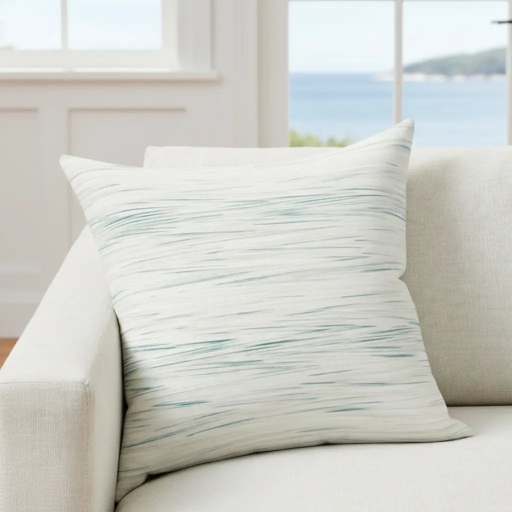 Coastal Breeze Square Pillow with blue watercolor stripes on pale linen-like fabric on a light sofa near a window.