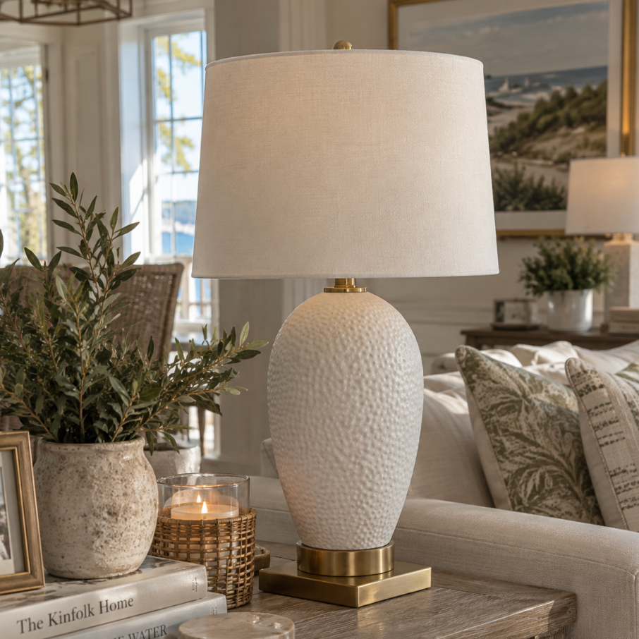 White textured ceramic table lamp with hammered detail, antique brass base, and linen drum shade on a coastal living room side table